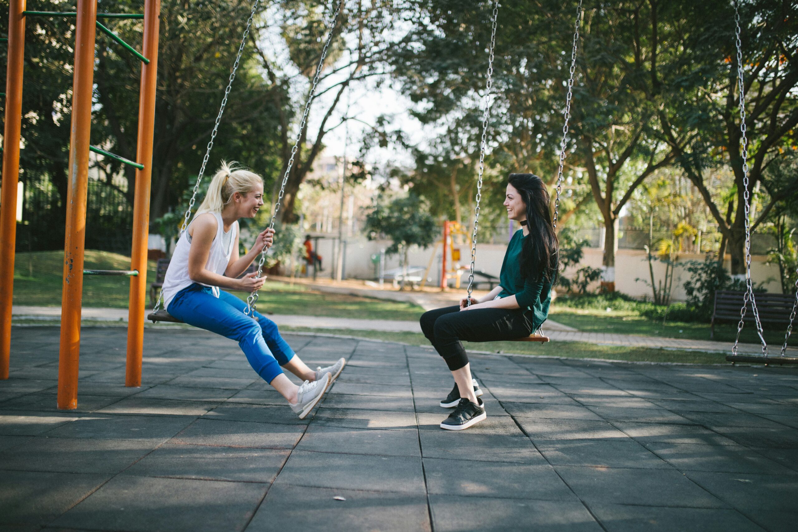 Two adult women talking while sitting on swings at a playground.