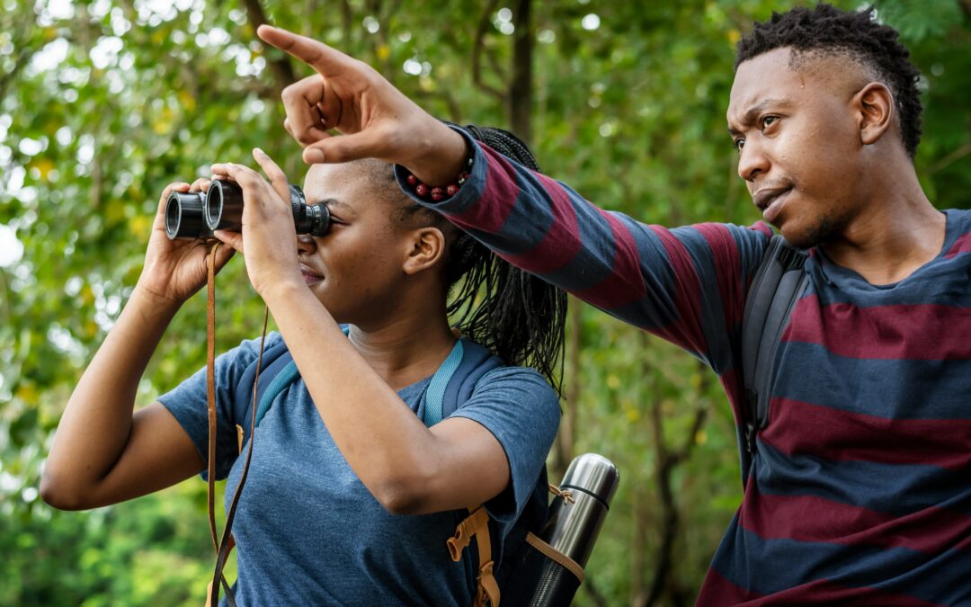 2 people in woods looking, one with binoculars, and pointing